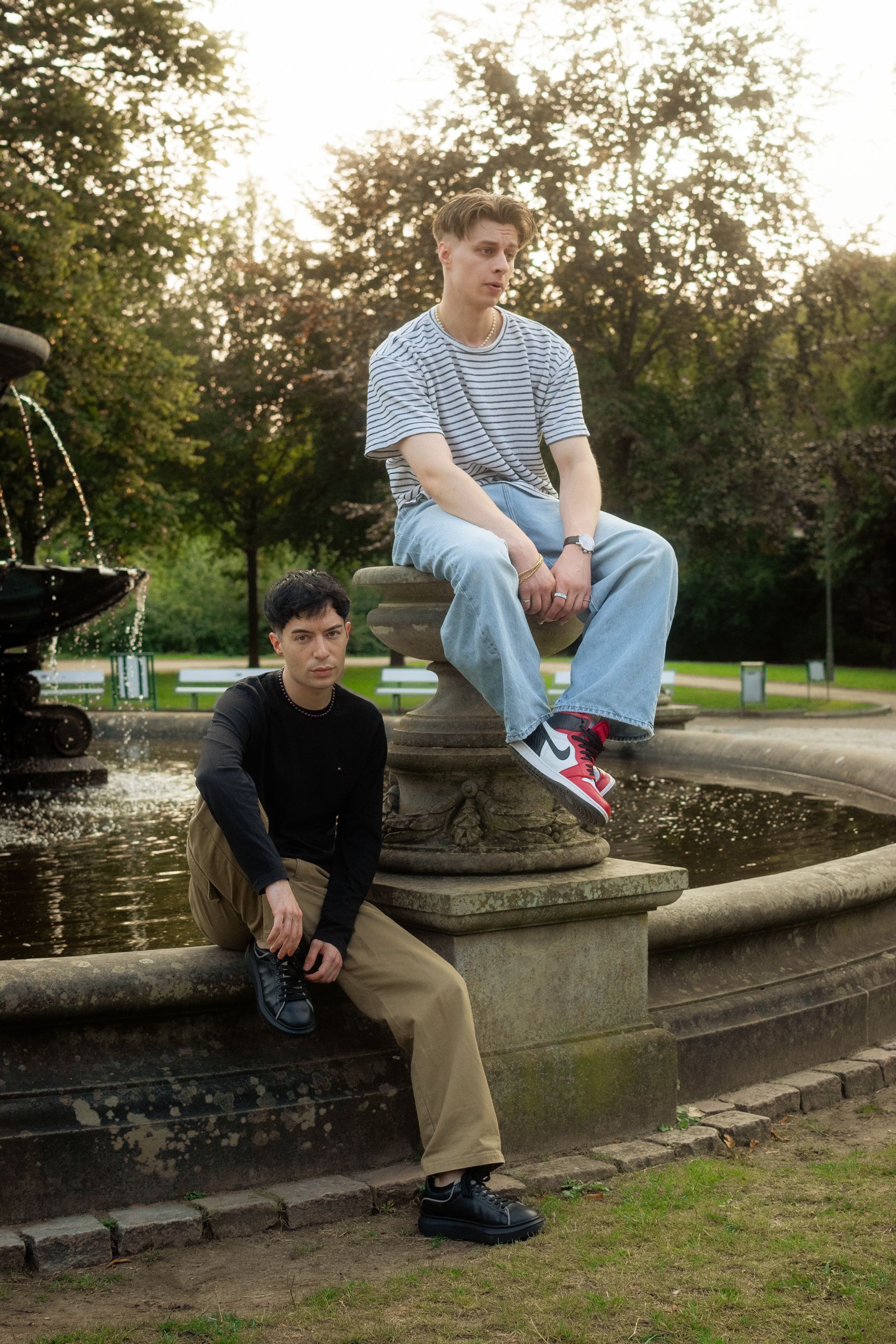 Two young adults in casual attire pose stylishly by a decorative fountain in a park.