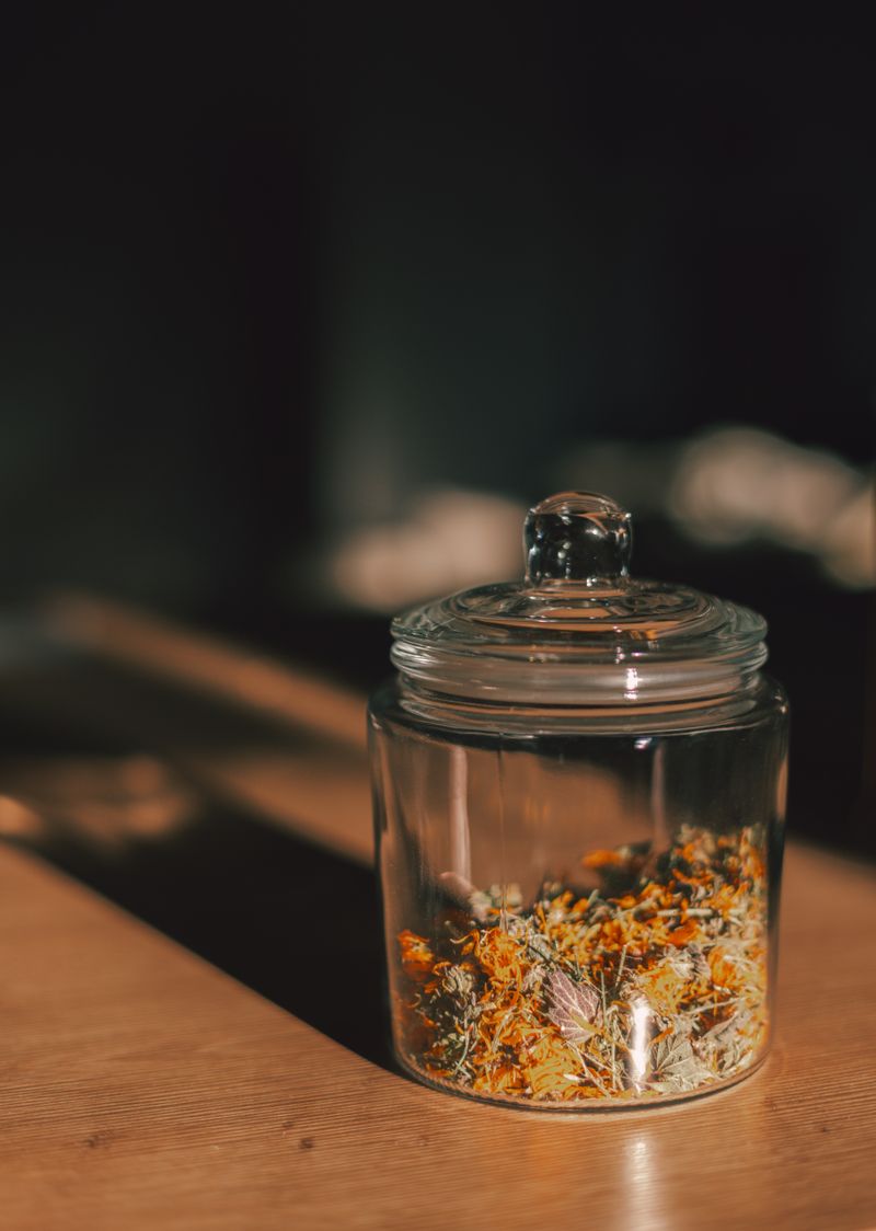 A glass storage jar of dried herbs and petals on a warm-lit wooden table. Photo by Raymond Petrik on Pexels.