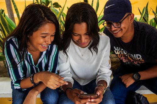 Free Three young friends in Barranquilla share a moment around a smartphone, smiling and bonding. Stock Photo