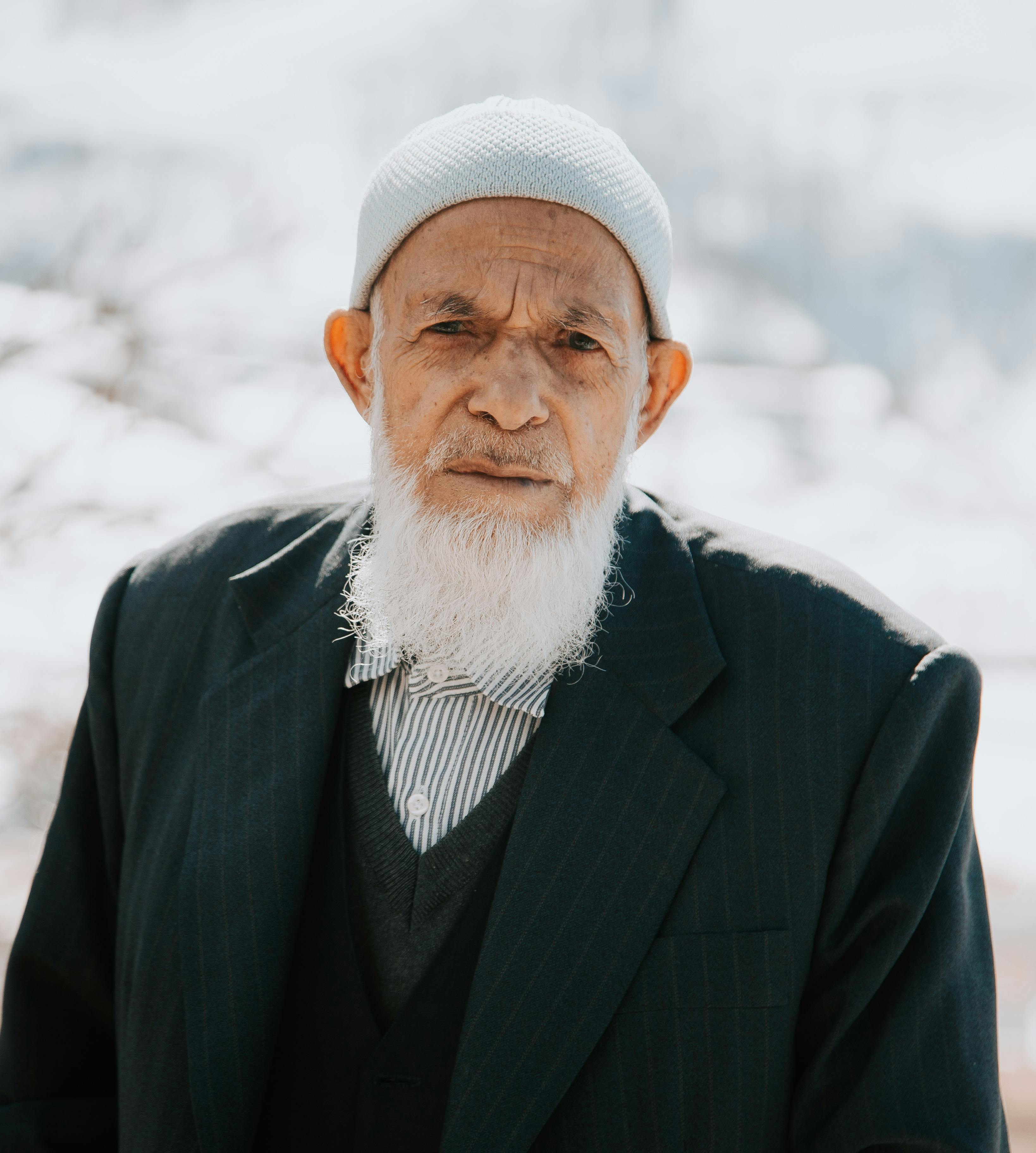Portrait of a senior man with a long beard wearing a suit and cap outdoors.
