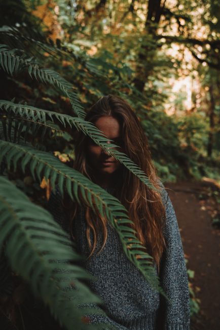 A woman in a sweater stands partially hidden by fern leaves in a forest.