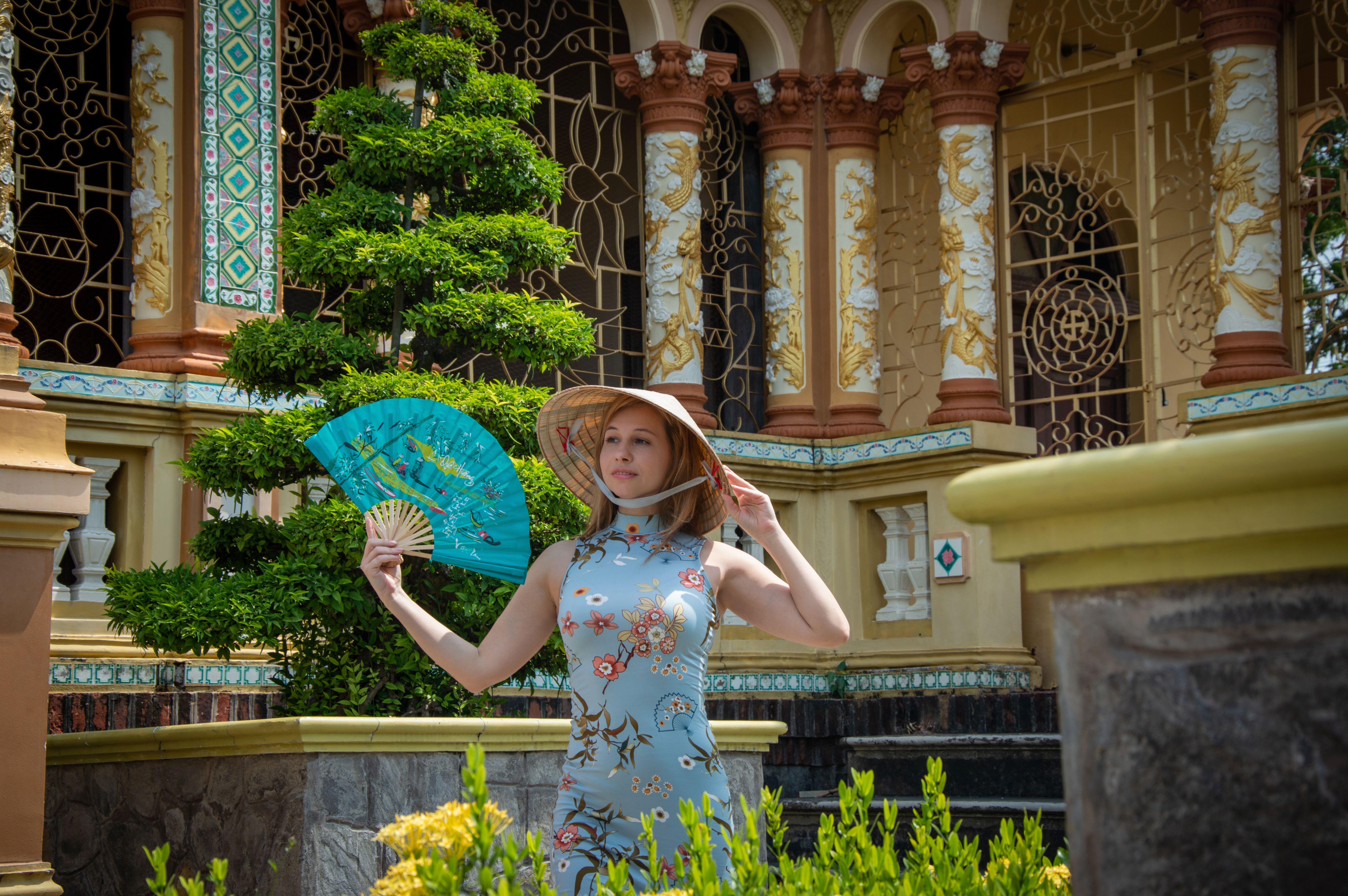 A woman poses with a fan in front of intricate Asian architecture, embodying cultural elegance.