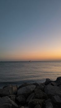 Tranquil sunset over the ocean with a distant boat on the horizon and rocky shoreline.