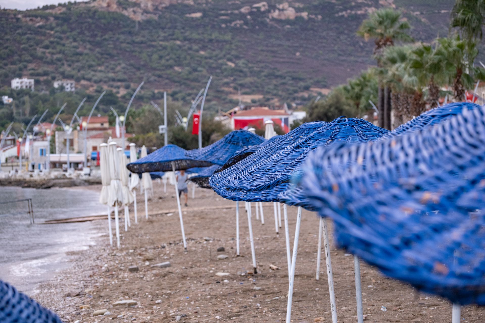 Turkish coastal scene in Foça İzmir featuring blue parasols along a sandy beach, under a cloudy sky.