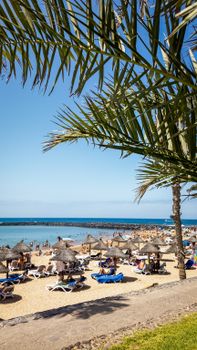 Busy beach scene at Playa de las Américas, Spain with people relaxing under parasols.