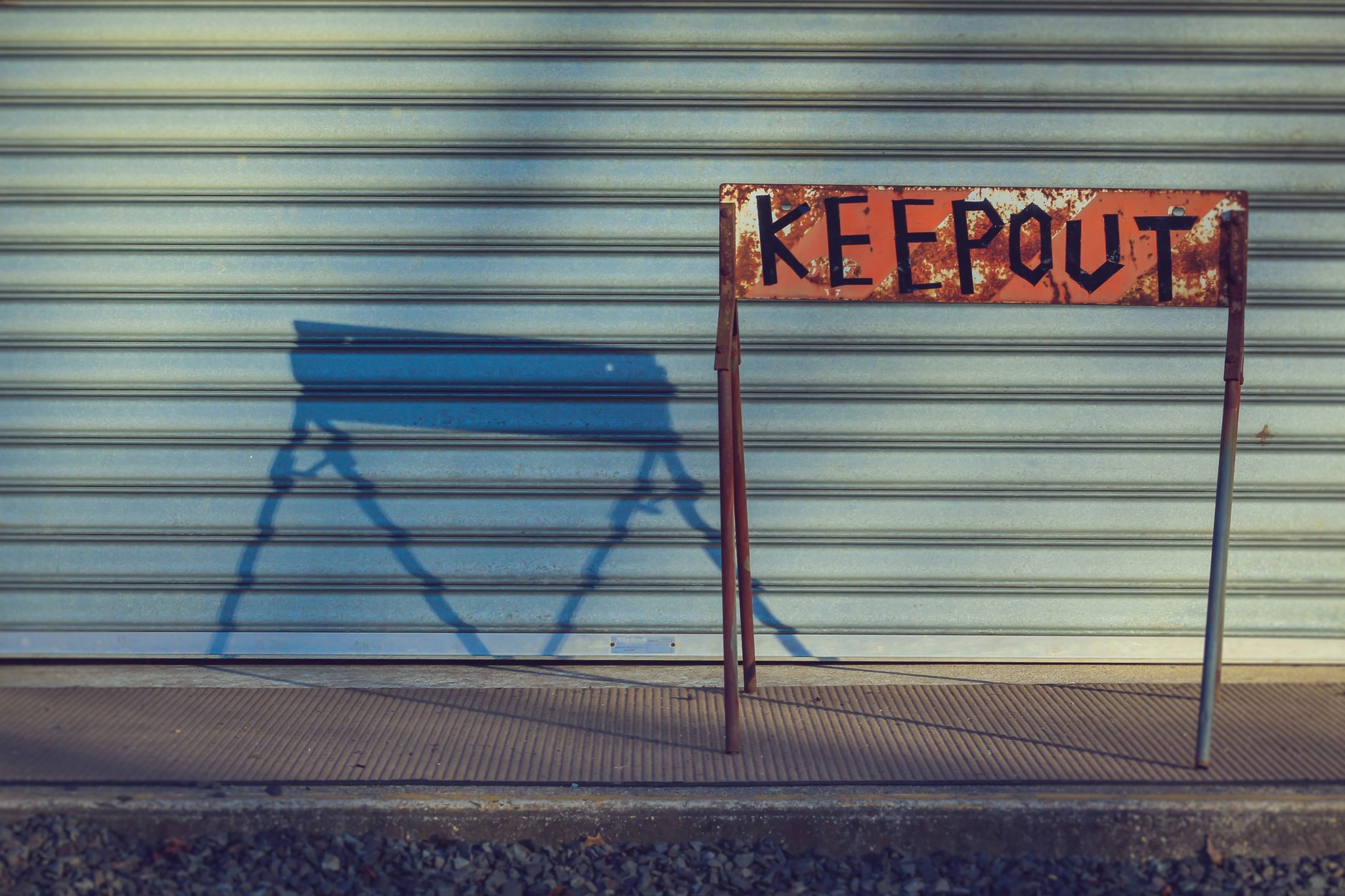 A weathered 'Keep Out' sign in front of a closed metal shutter, casting a shadow.