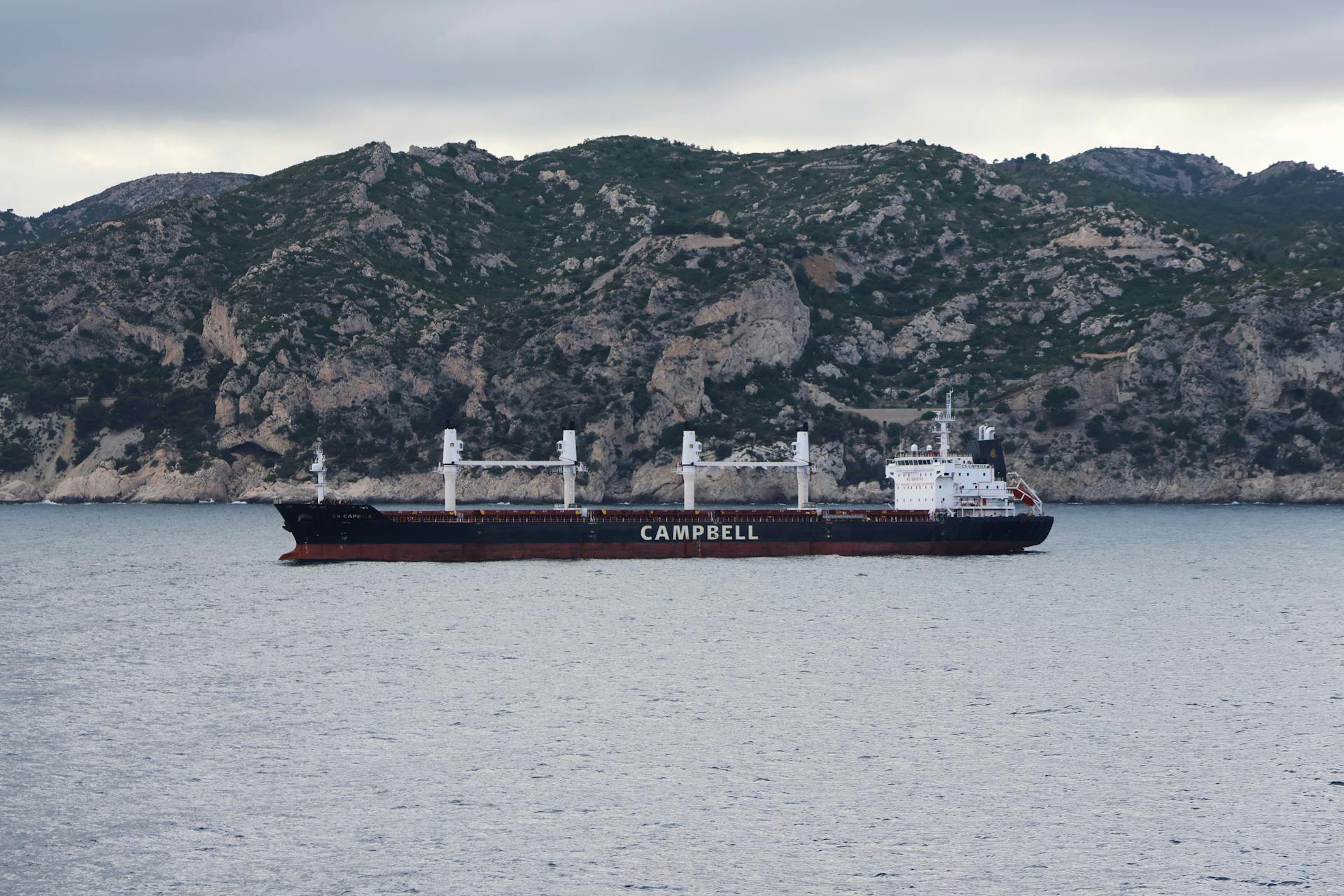 A black cargo ship labeled 'Campbell' sailing near rocky cliffs under a cloudy sky.