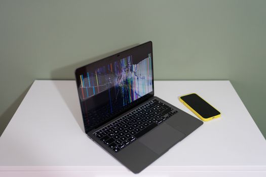 A damaged laptop screen with colorful lines and a yellow smartphone on a white desk.
