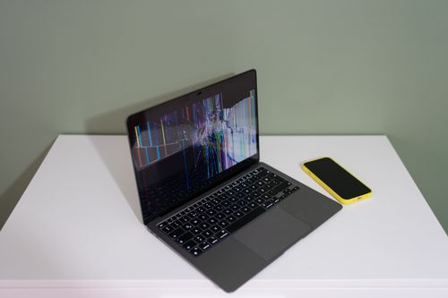 Free A damaged laptop screen with colorful lines and a yellow smartphone on a white desk. Stock Photo