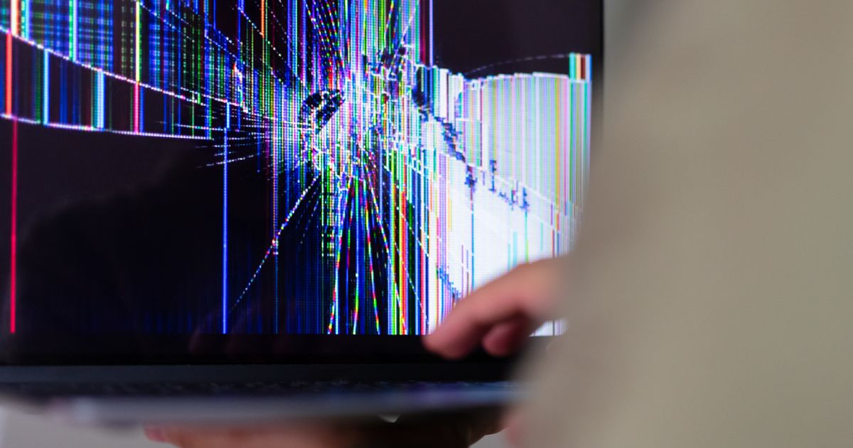 A laptop screen shattered into coloured vertical lines and spreading cracks, held open in one hand