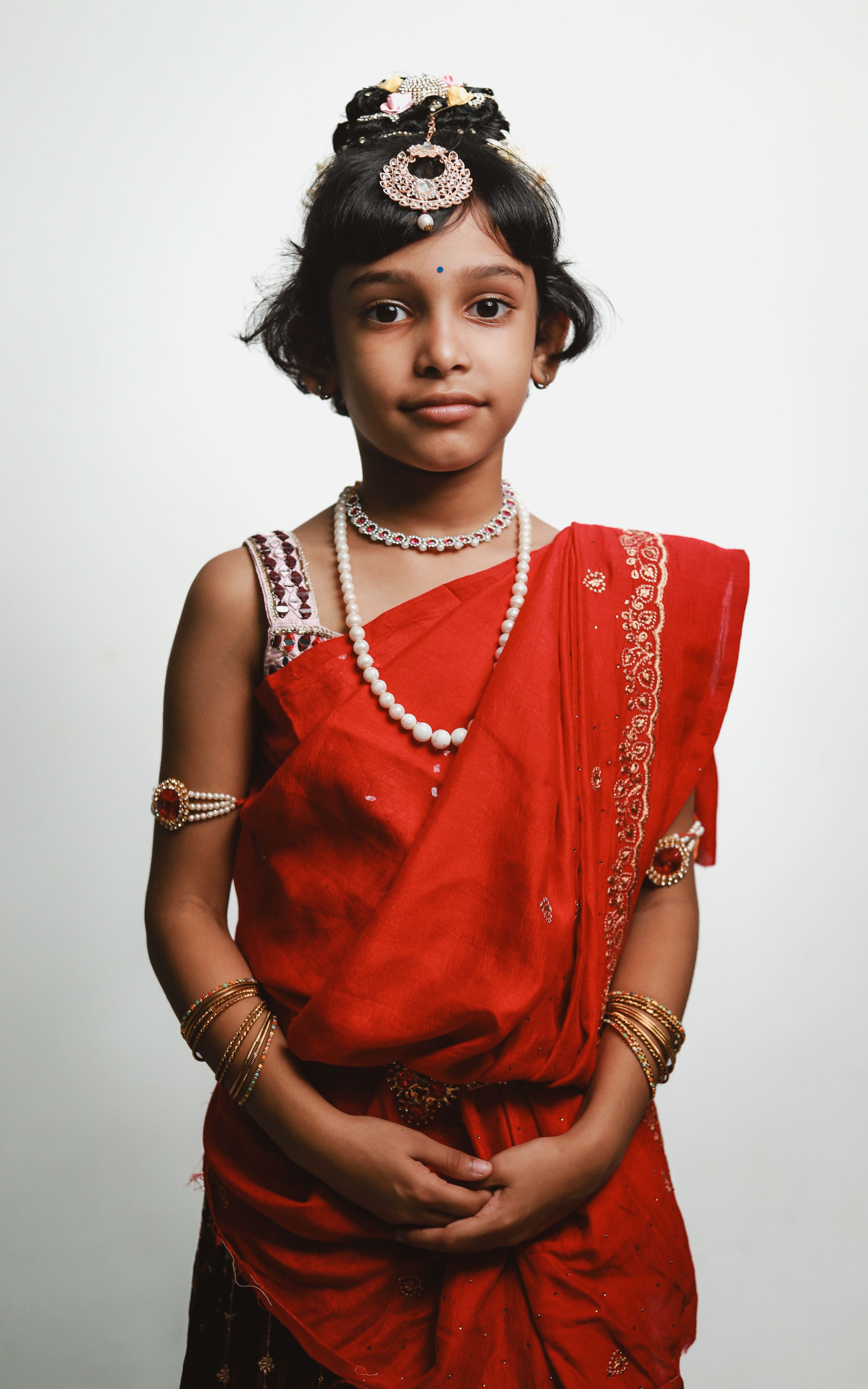 Young girl adorned in red traditional Indian attire and jewelry, posing gracefully.