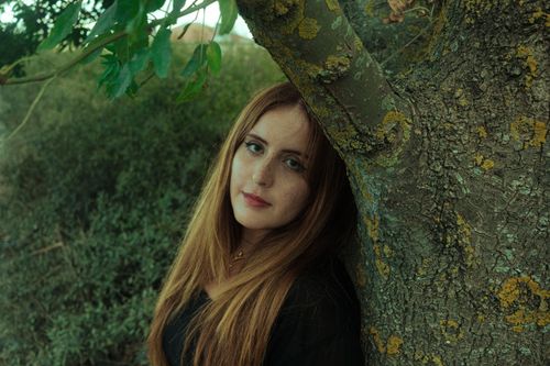 Free Woman with long hair leaning against a tree, in a natural outdoor setting for reflective mood. Stock Photo