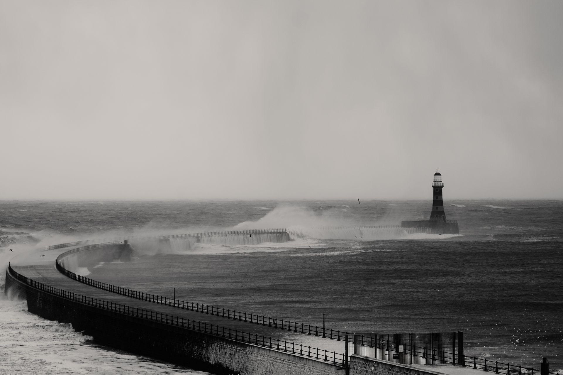 A lighthouse stands resilient amid crashing waves on a stormy black and white seascape.