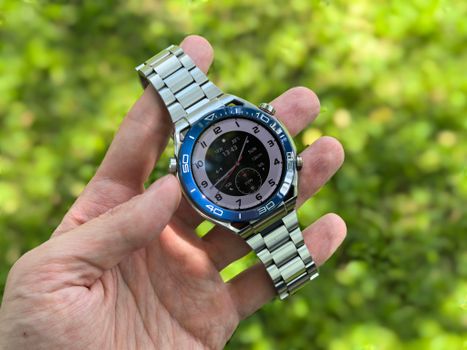Close-up of a modern silver wristwatch held in hand with green background outdoors.