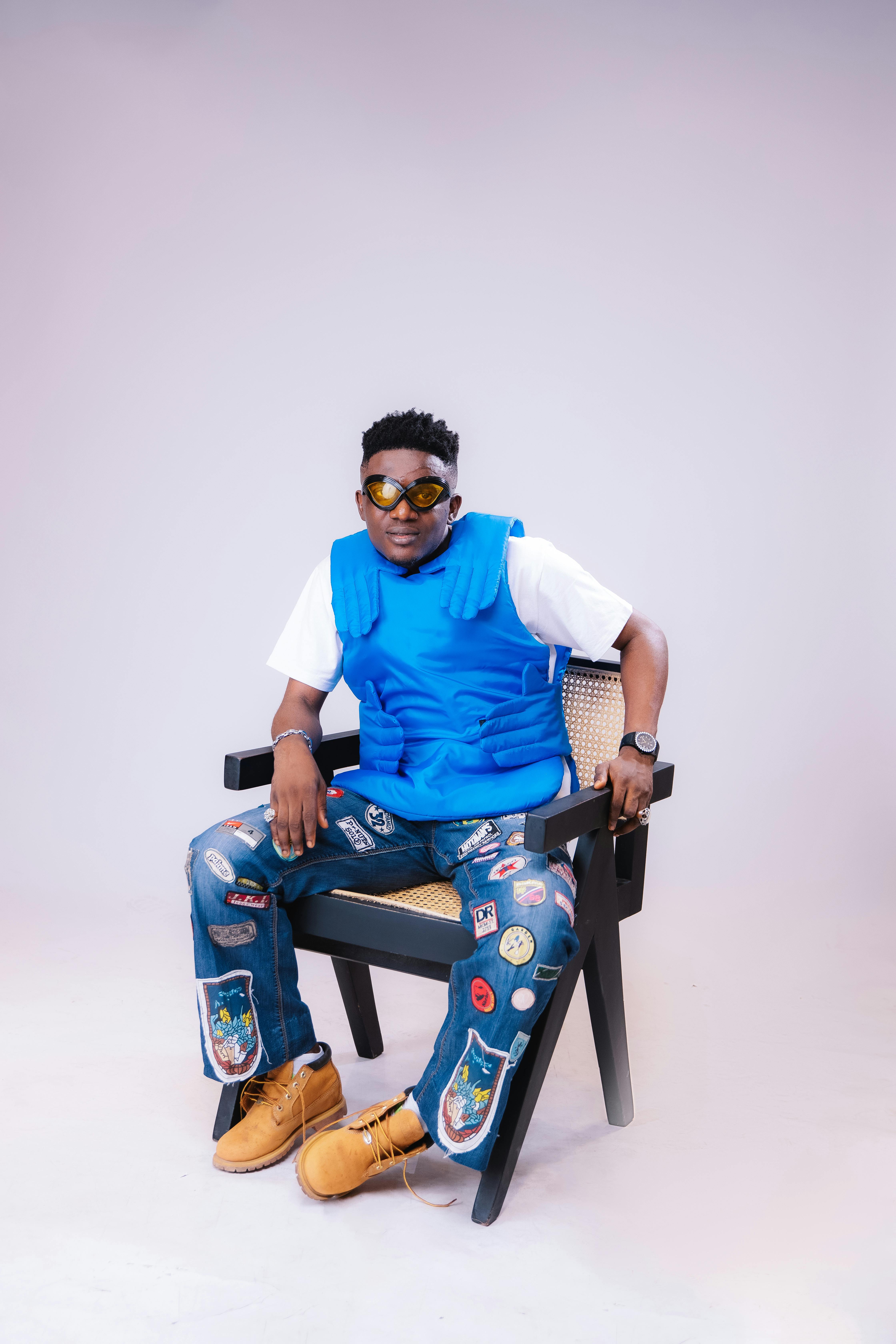 Young man wearing stylish clothing with sunglasses, seated confidently in a studio setting.