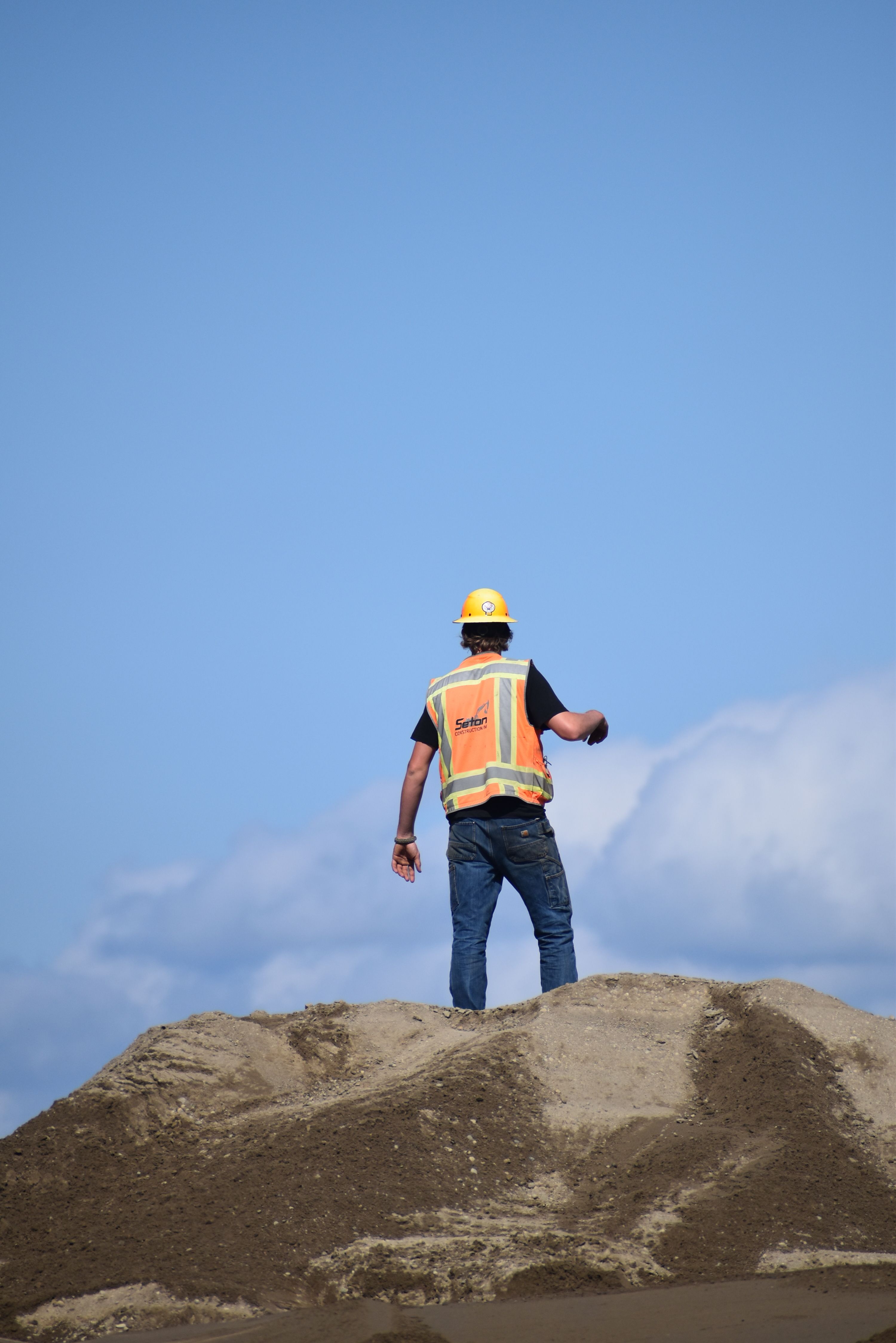 Construction worker in hard hat and vest surveying a sandy mound against a clear sky.