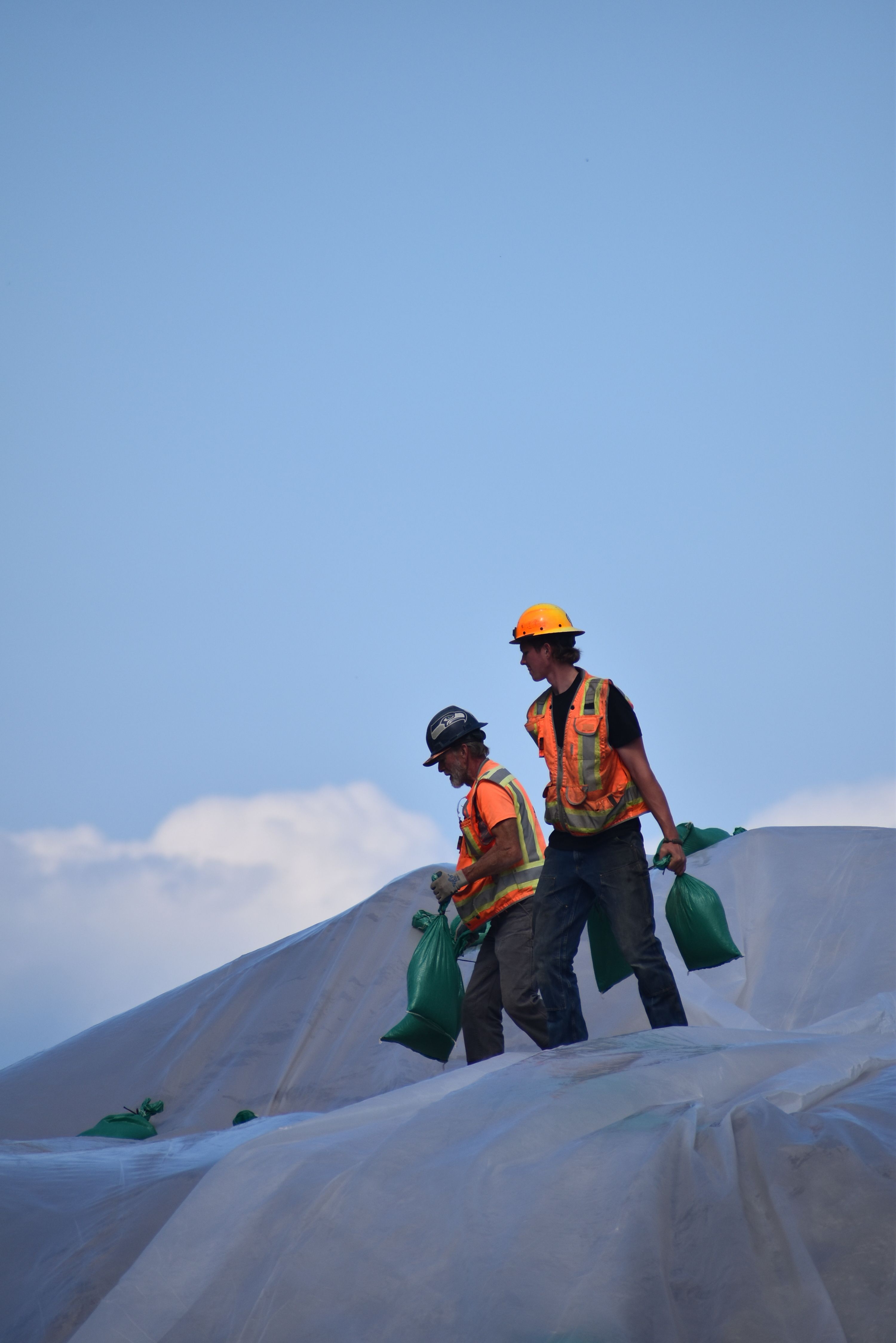 Two construction workers securing plastic sheeting with sandbags on a bright sunny day.