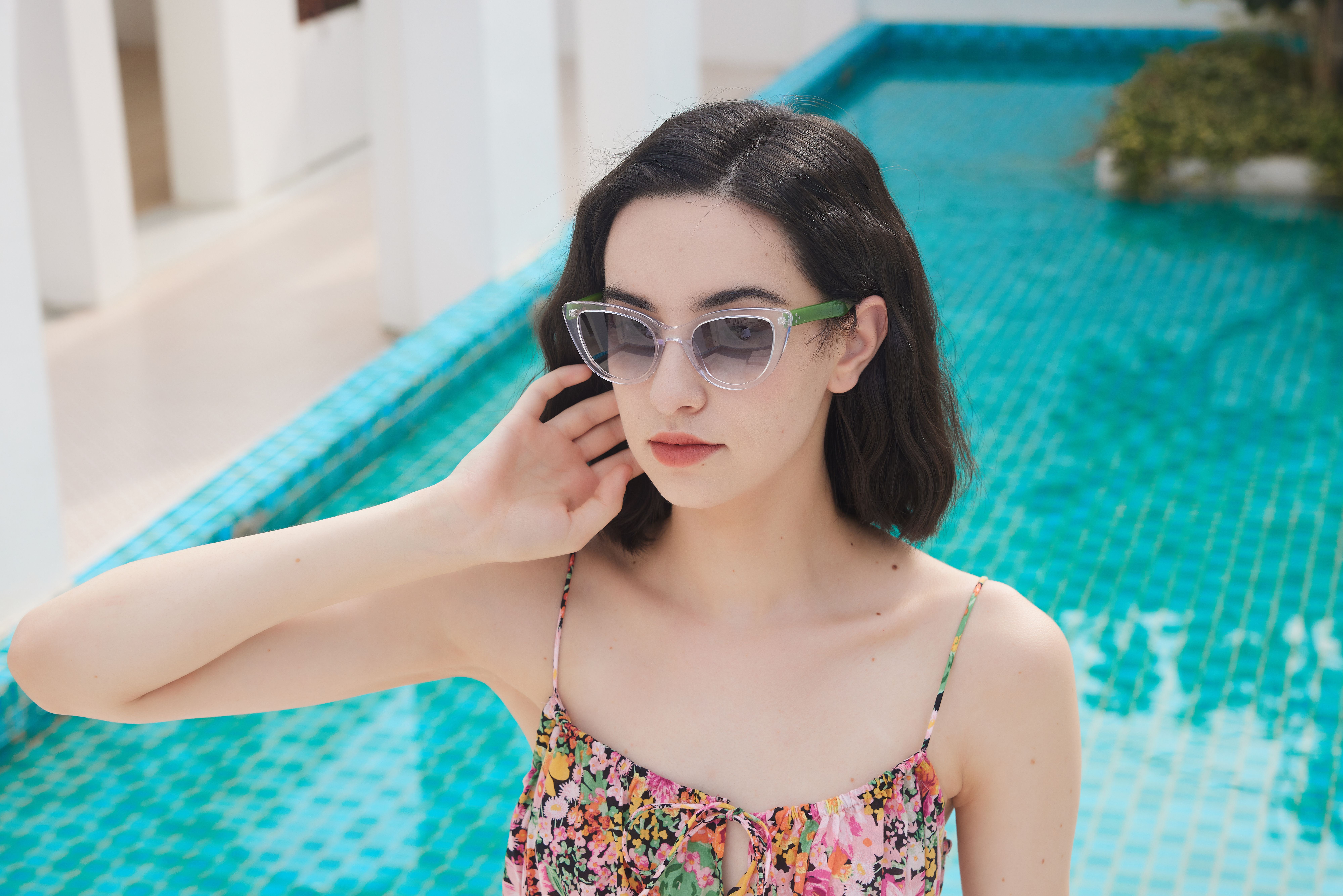 Stylish woman in sunglasses and floral dress posing by a pool on a sunny day.