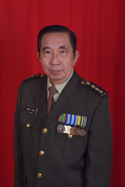 Distinguished older male military officer in uniform with medals, set against a red backdrop.