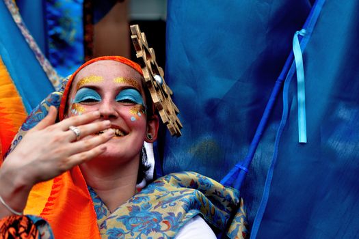 A joyful woman with artistic makeup and a vibrant headdress, smiling amid bright fabrics.