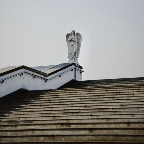 Free A peaceful angel statue stands atop a rooftop staircase, offering a glimpse of tranquility against the sky. Stock Photo