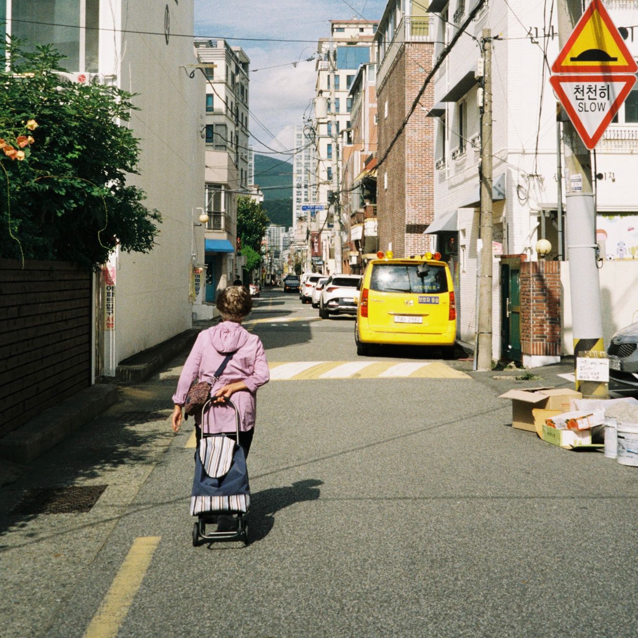 Elderly woman pulling a cart down a bustling city alley with parked cars and buildings.