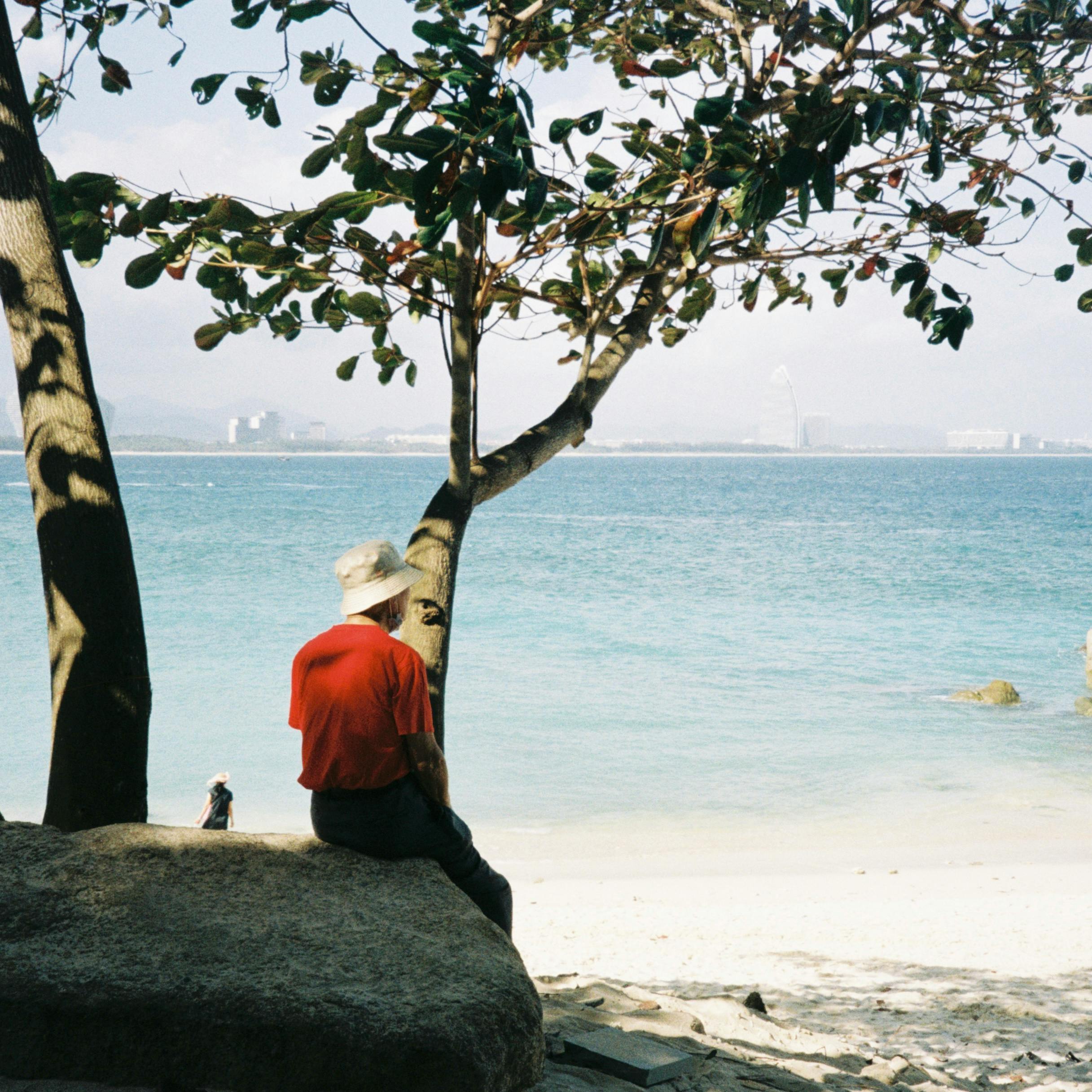 Elderly man in hat enjoys a peaceful moment by the seaside under a tree.
