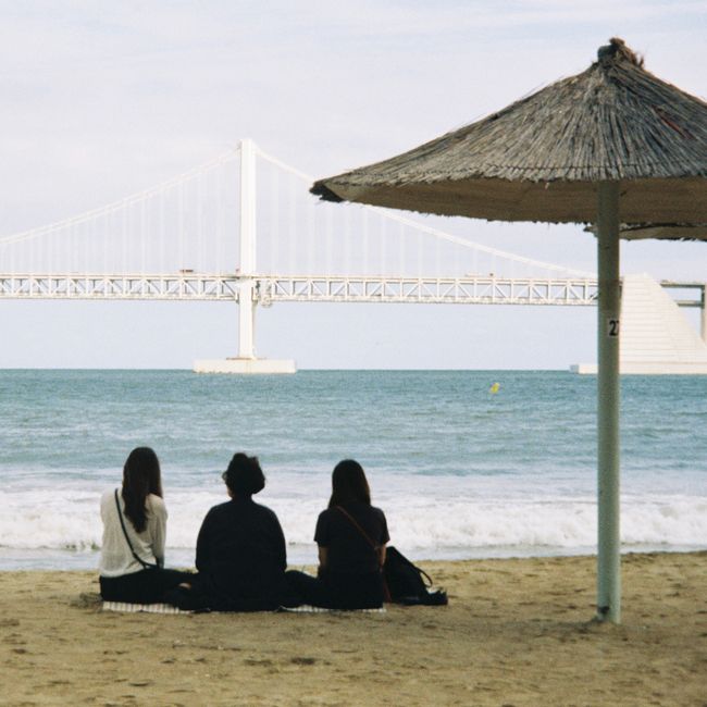 Three people enjoying a peaceful moment on a beach facing the iconic Gwangan Bridge.