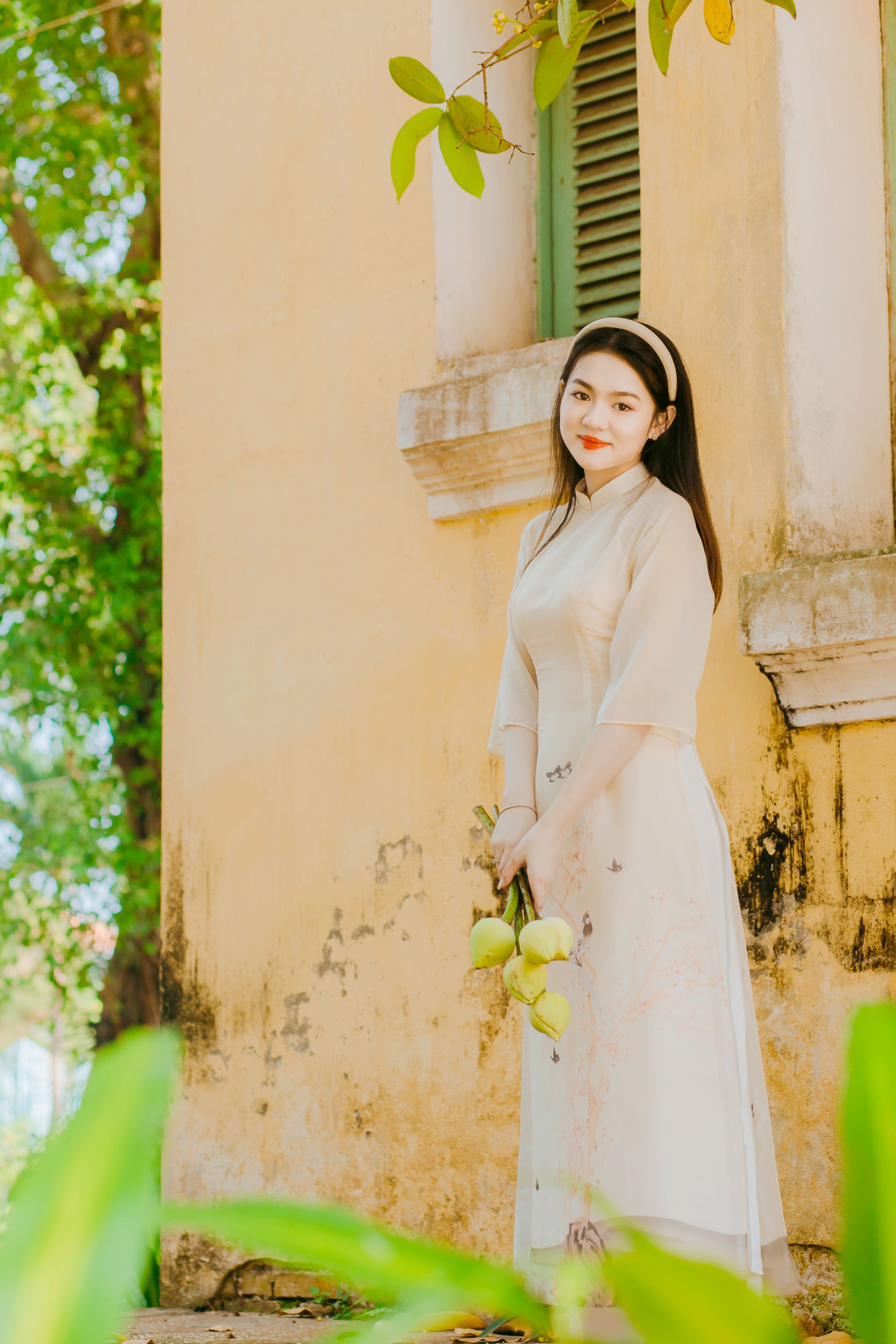 Young woman in an ao dai dress holding flowers near a rustic yellow wall, embodying elegance and tradition.