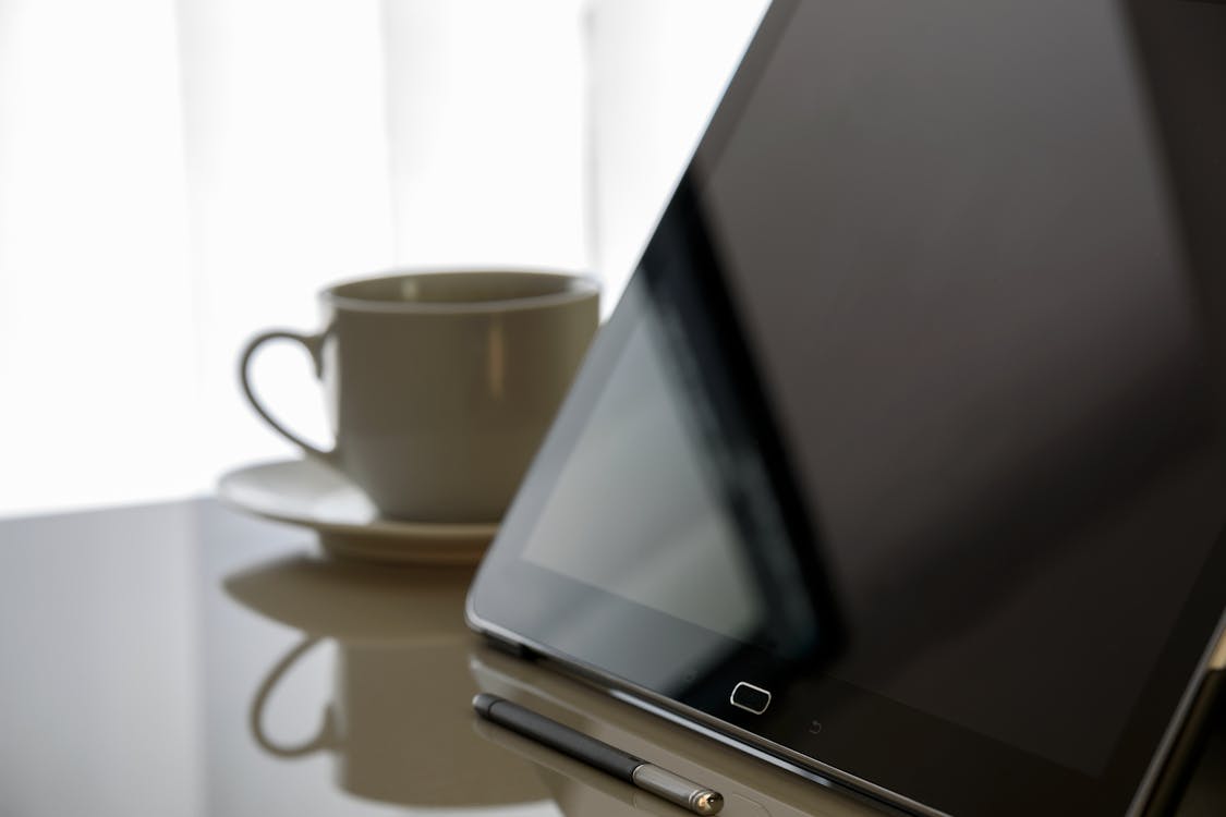 бесплатная Черный планшетный компьютер рядом с чашкой с блюдцем на столе Стоковое фото
