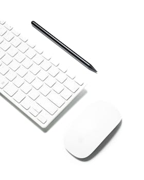 A clean and minimalist workspace featuring a wireless keyboard, mouse, and stylus on a white background.