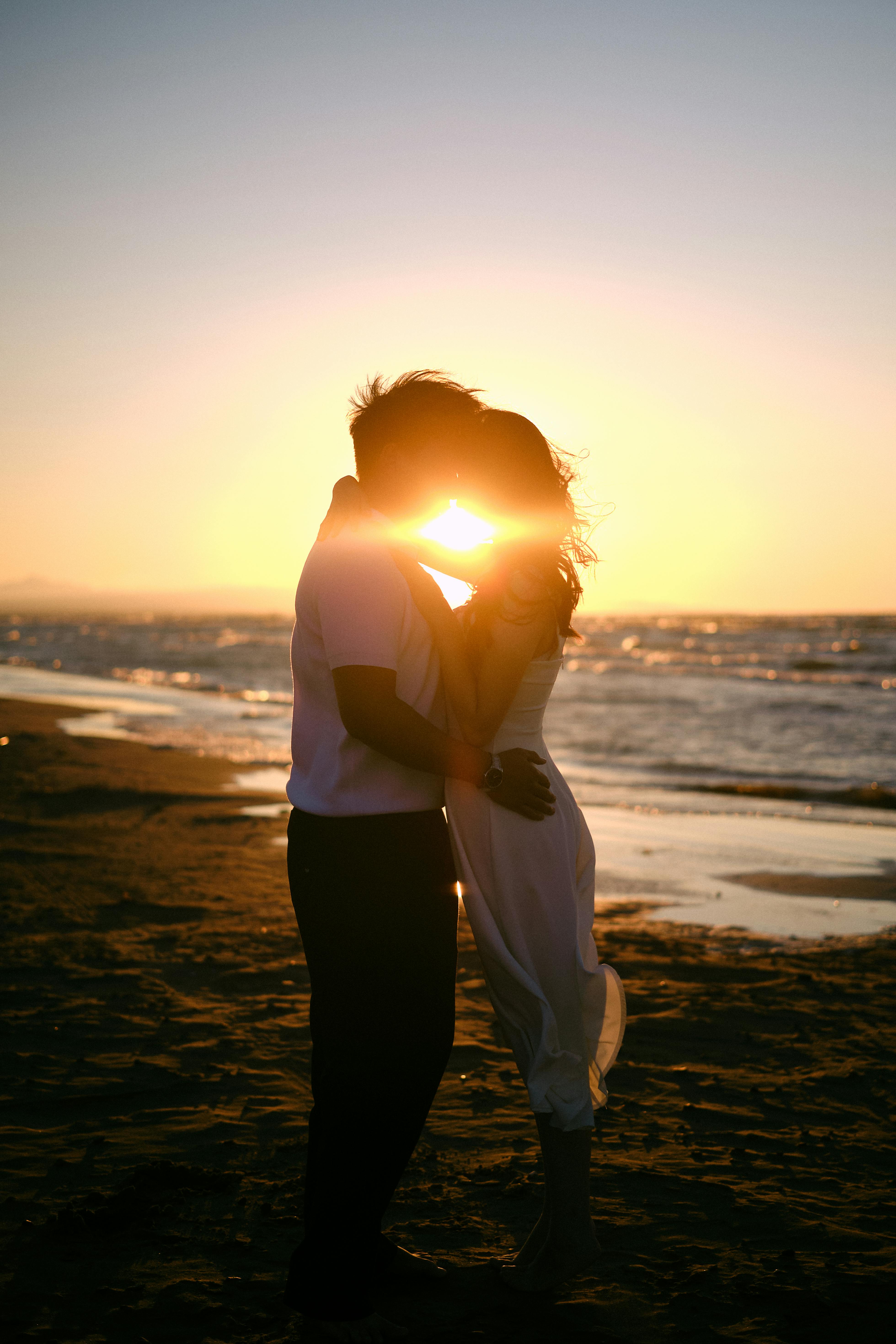 A couple embraces on the beach at sunset, creating a romantic silhouette against the sunlit ocean waves.
