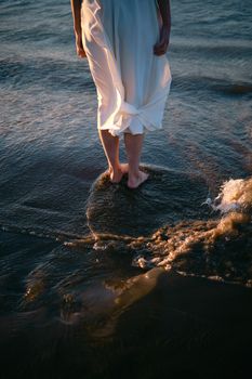 A woman in a white dress walks barefoot along the shore at sunset, capturing tranquility and freedom.