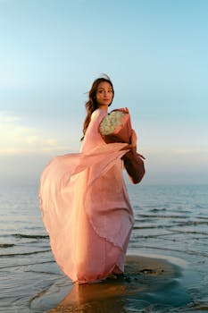 A woman in a flowing pink dress holding flowers, standing on a sandy beach at sunset.