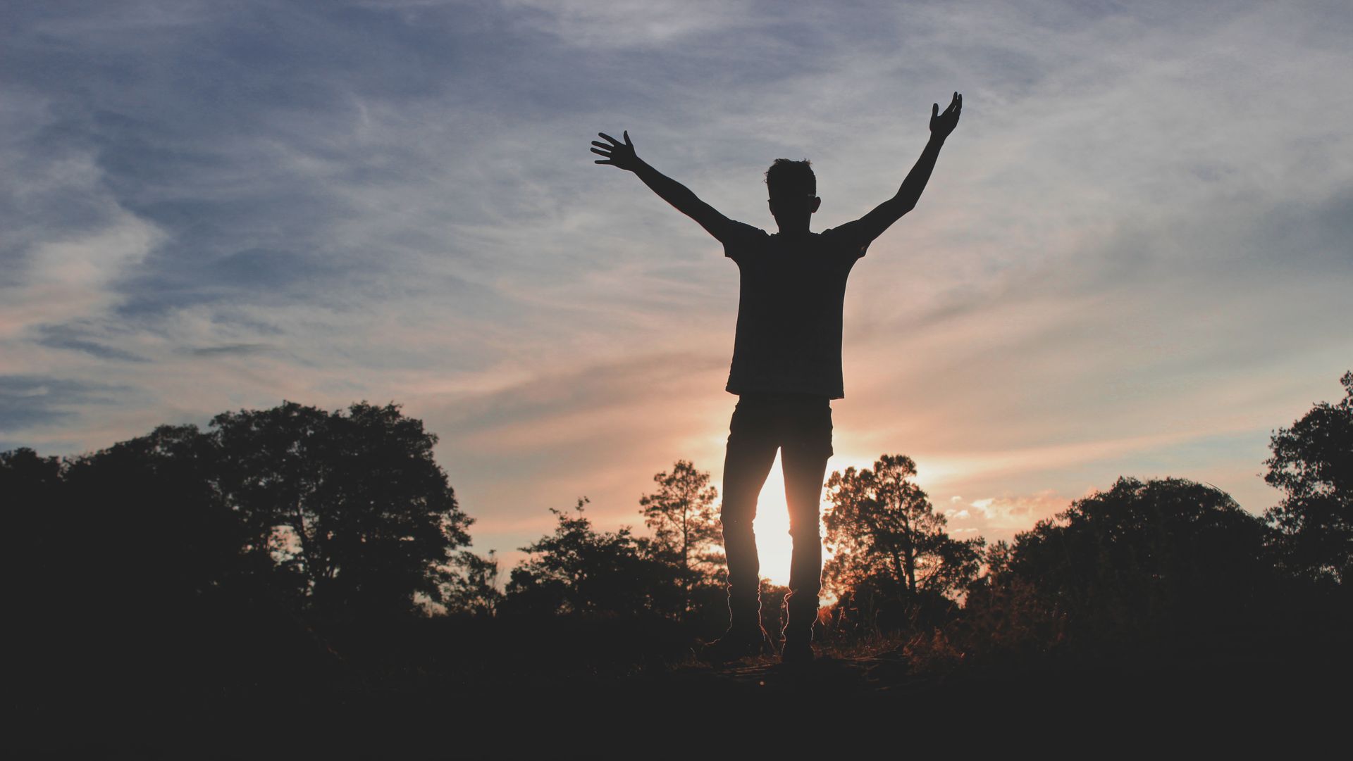 Silhouette of a person raising arms with a dramatic sunset backdrop and trees.