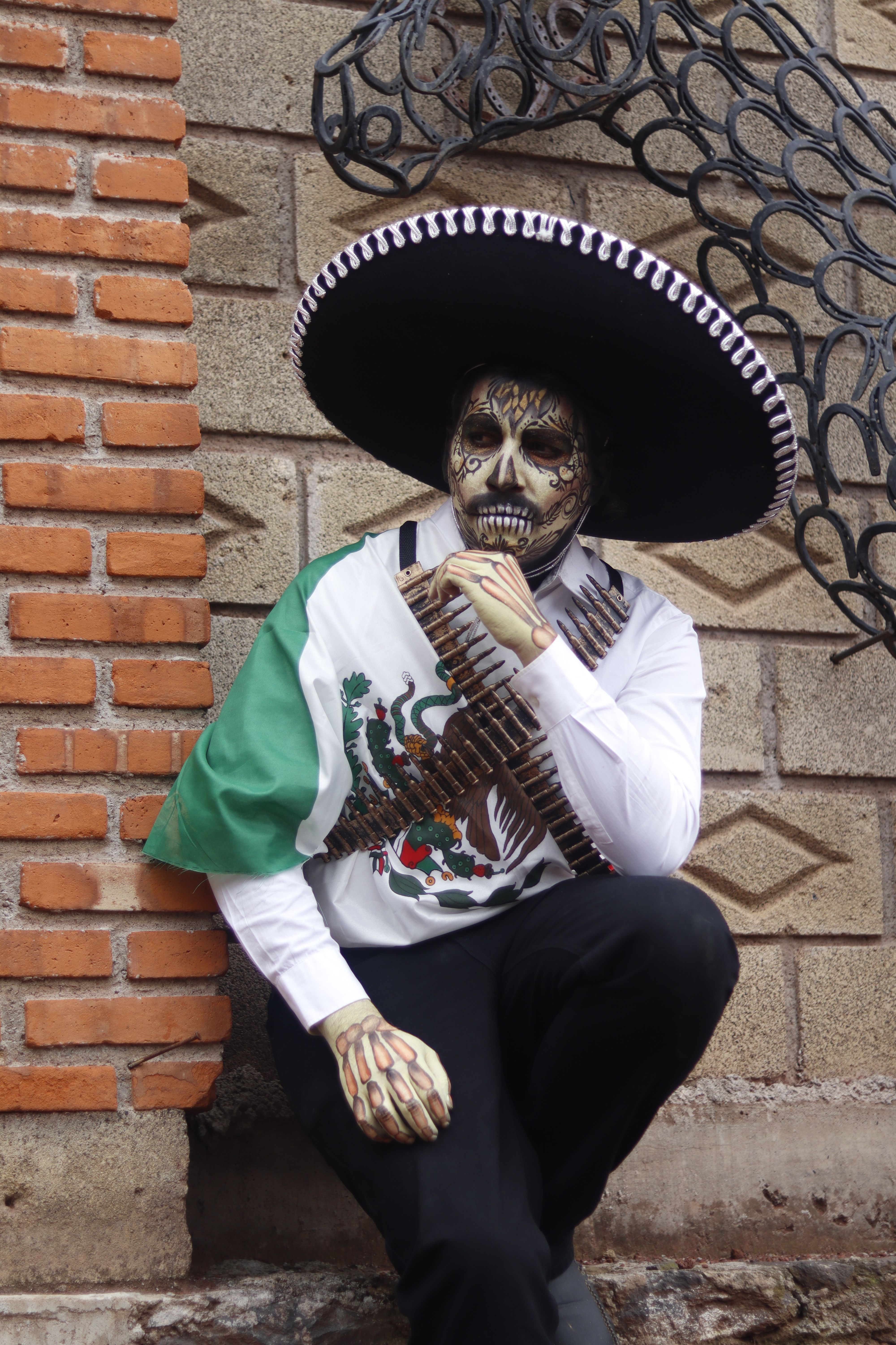 A person dressed in Day of the Dead attire with face paint, sitting by a brick wall.