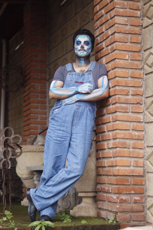 Free A man wearing blue overalls and skeleton face paint leans against a brick wall, showcasing creative makeup art. Stock Photo