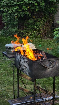 An outdoor barbecue grill with bright flames and a cooking pot in a lush garden setting.