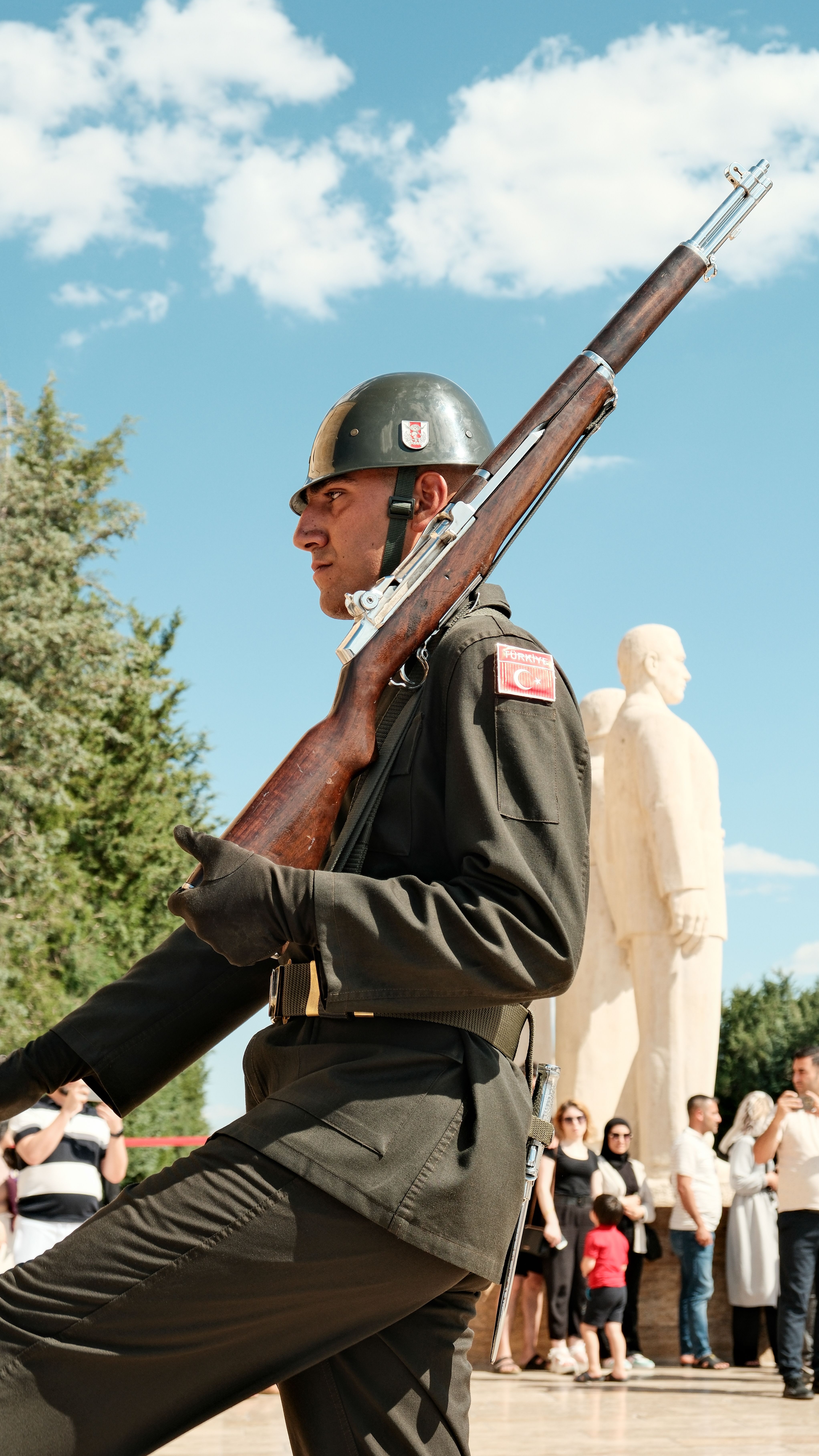 A soldier in ceremonial uniform marches before a statue on a sunny day with a crowd in the background.