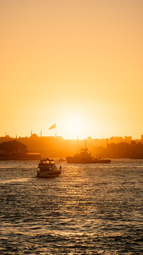 Free A serene view of boats sailing during a vibrant sunset over a coastal cityscape. Stock Photo
