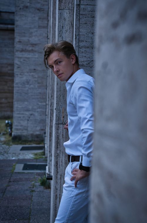 Free Stylish male model in white outfit leaning against a wall in urban Berlin, captured during twilight. Stock Photo