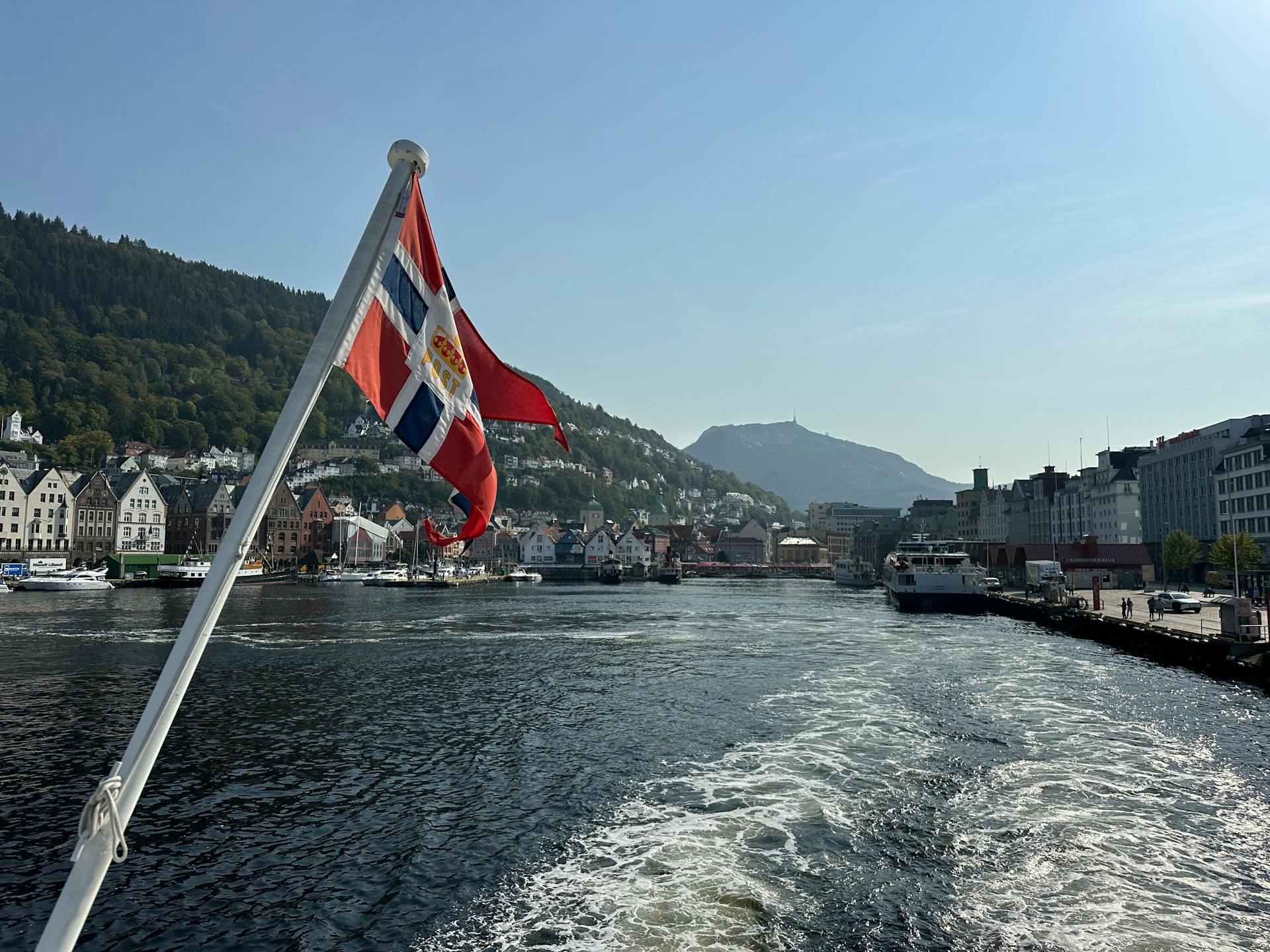 Norwegian flag waves over scenic Bergen harbor with iconic Bryggen buildings in the background.