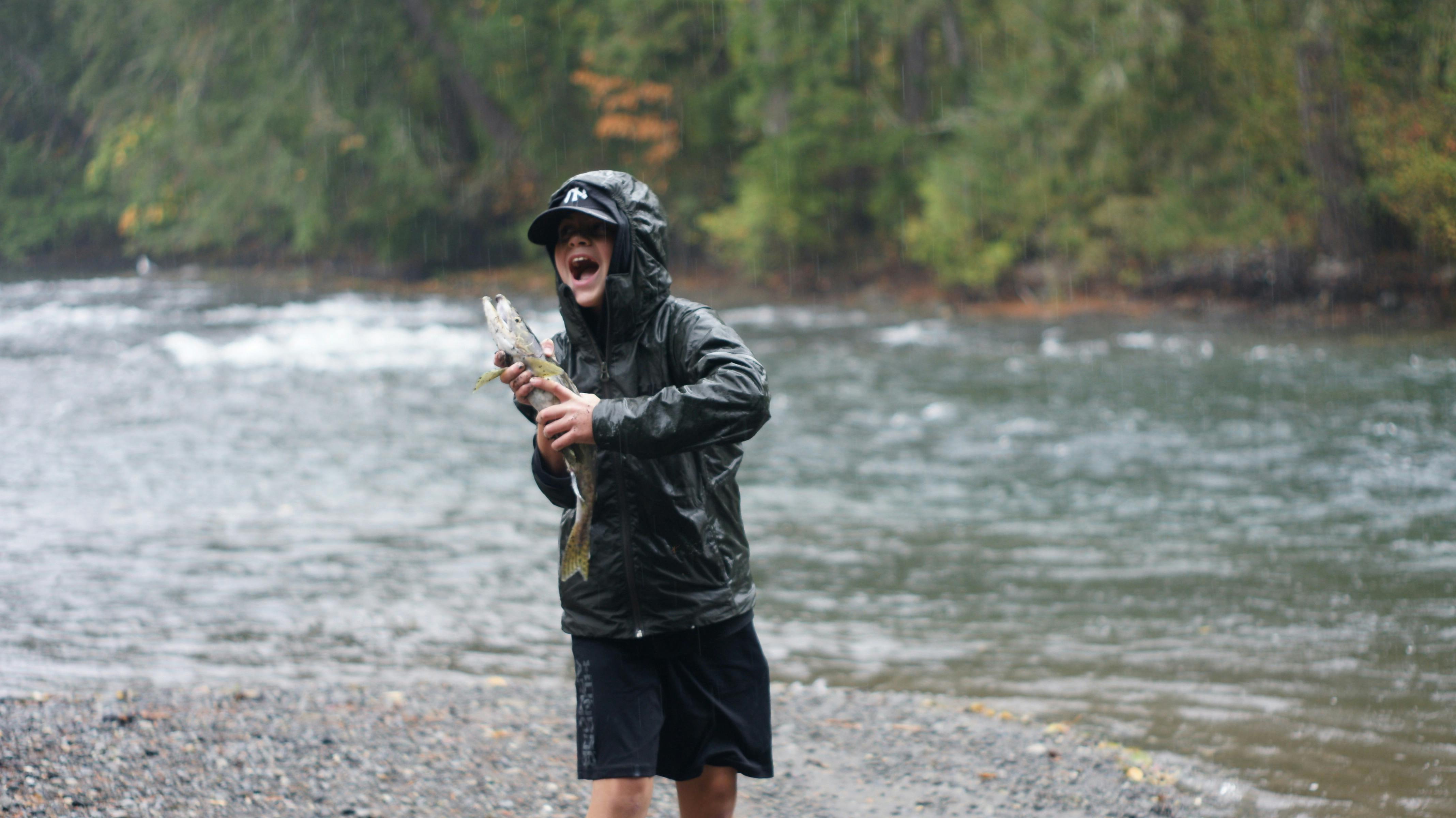 Boy celebrates catching a fish on a rainy day by the riverbank.