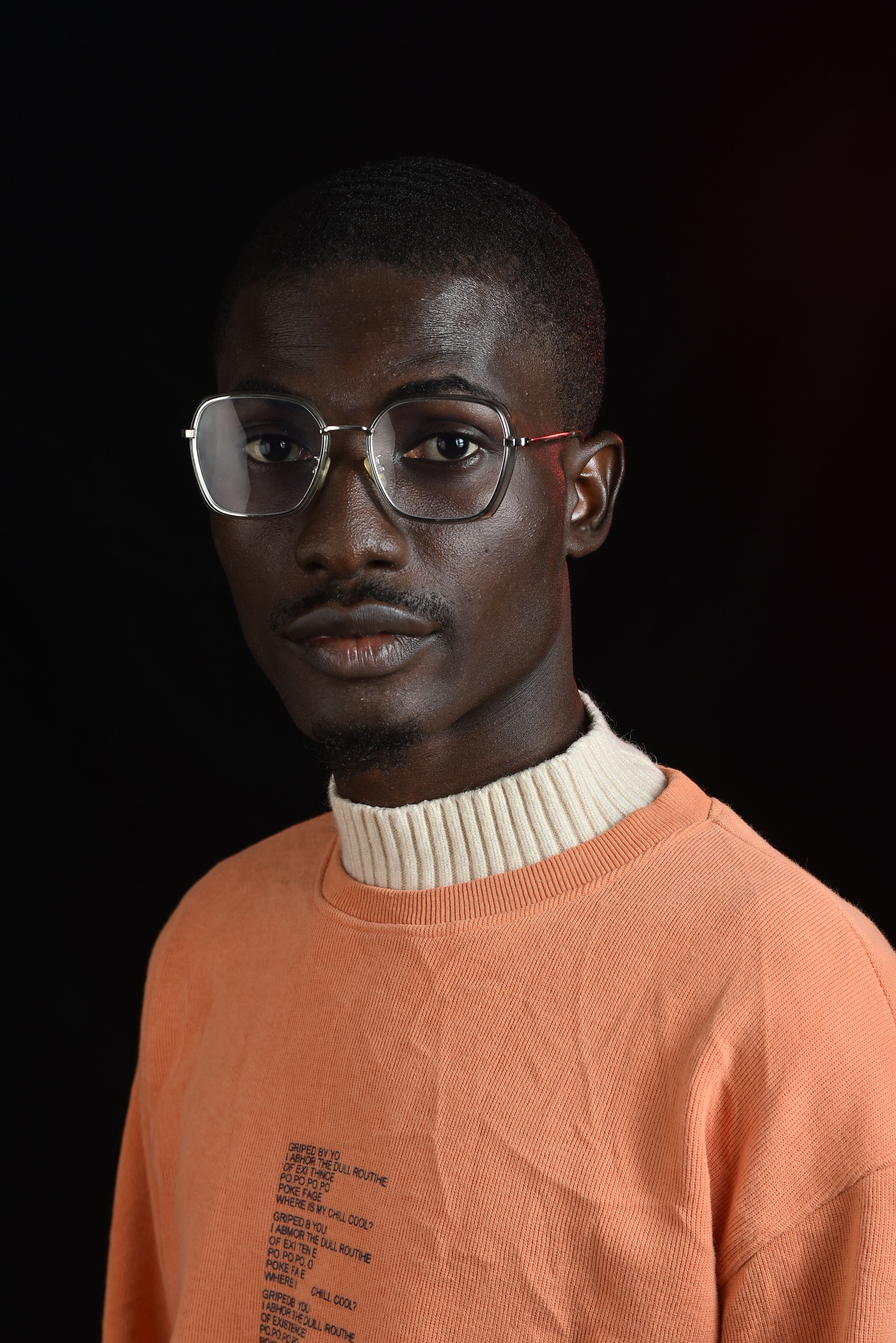 Stylish young man in glasses and orange sweater posing in a studio portrait with a black background.