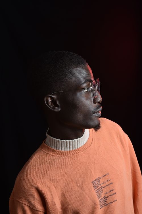 Free A young man in glasses looks away, wearing an orange sweater against a dark background. Stock Photo
