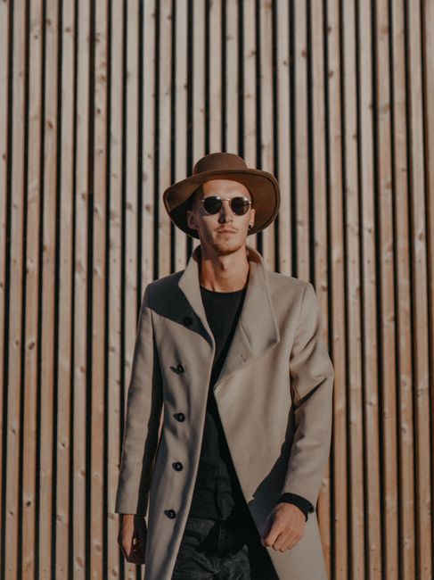 Fashionable man with a coat and hat poses against a wooden wall in warm sunlight.