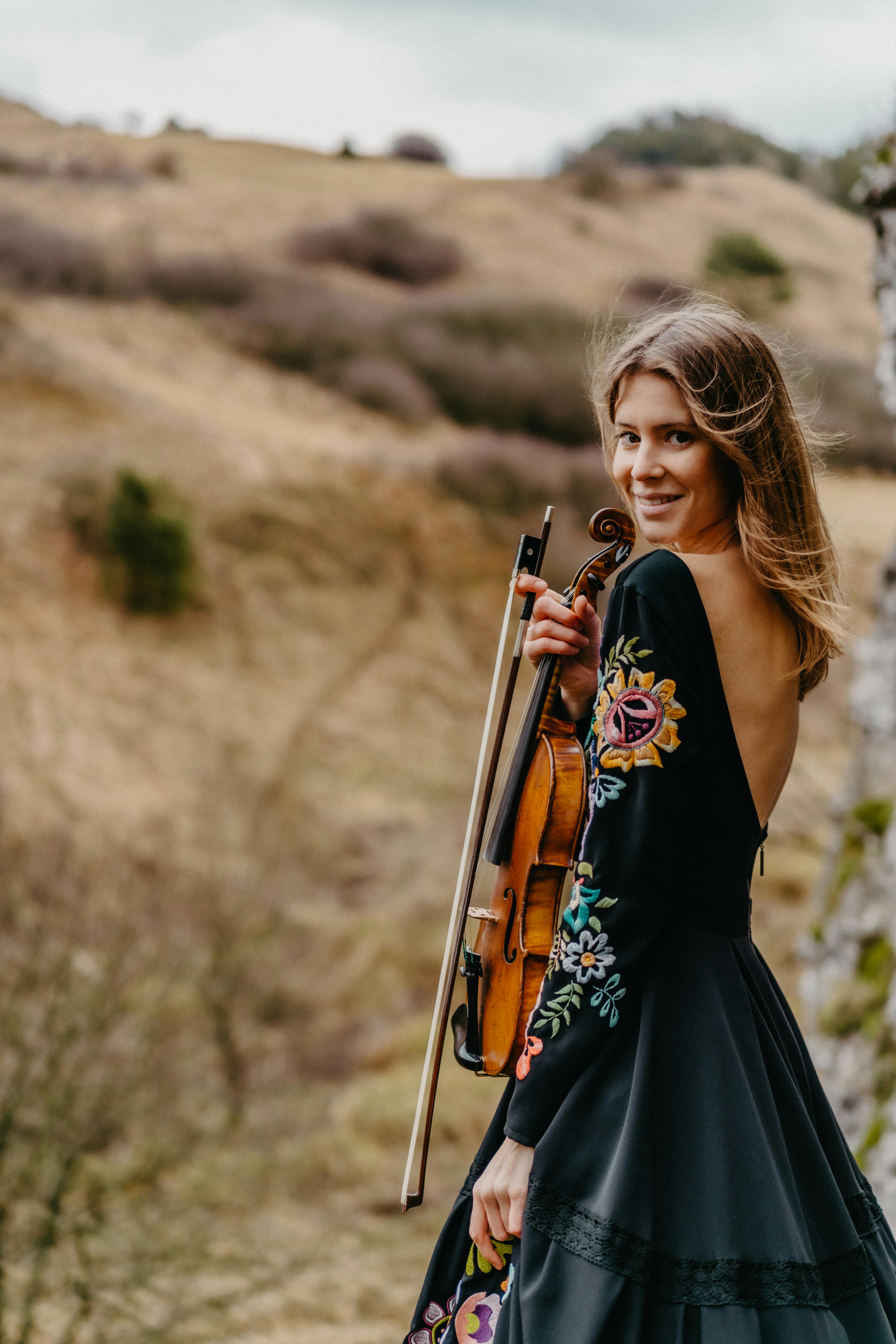 A young woman in a black floral dress plays violin on a scenic hillside.