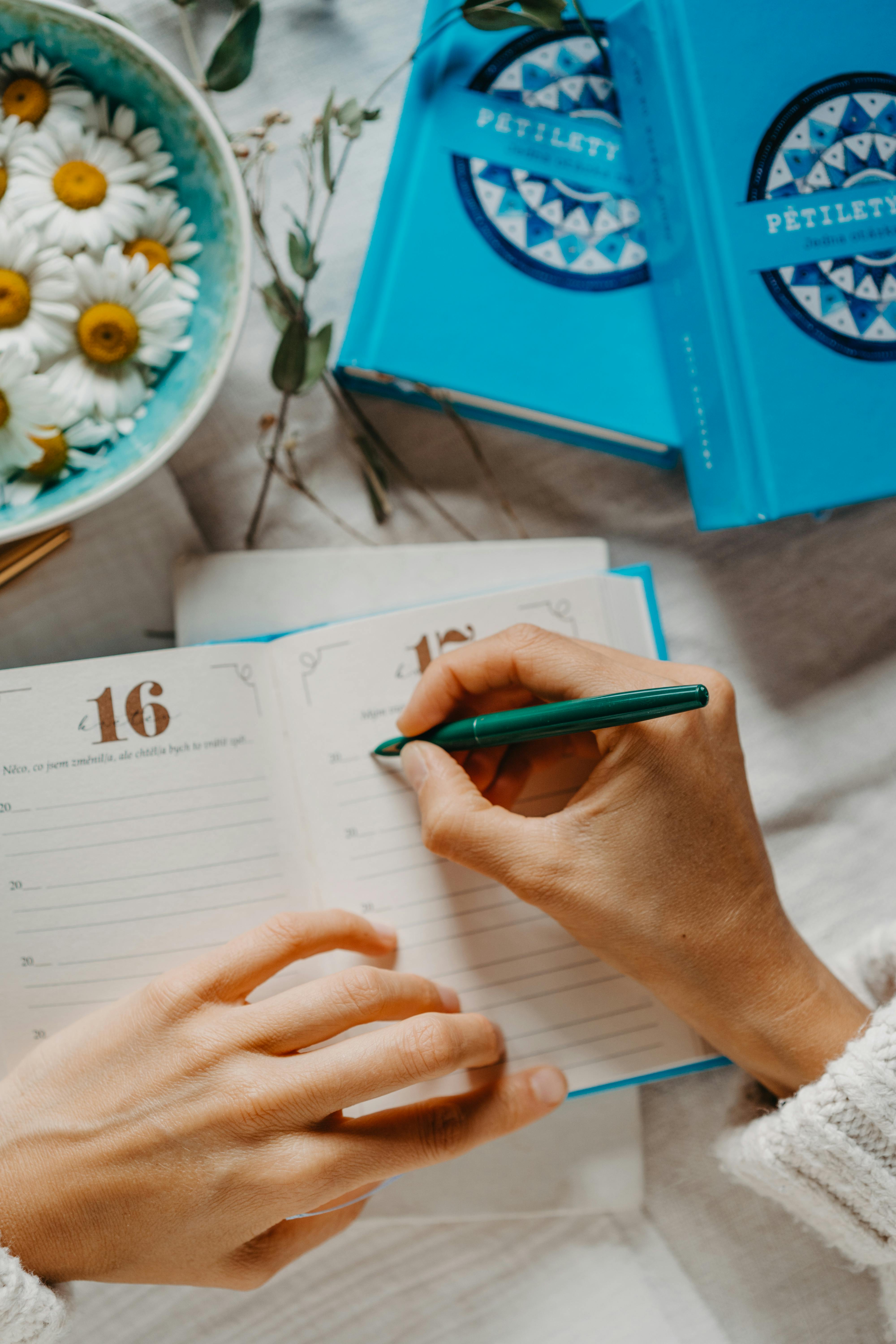 A woman writes in a blue journal, surrounded by daisies and books, in a cozy setting.