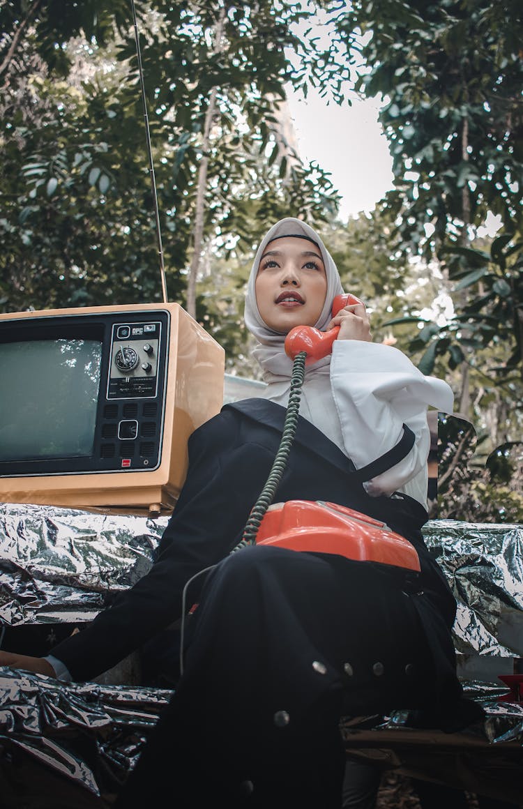 Low Angle Photo Of Muslim Woman In A Hijab Sitting On A Bench Posing With A Rotary Phone And An Old Television Set Beside Her