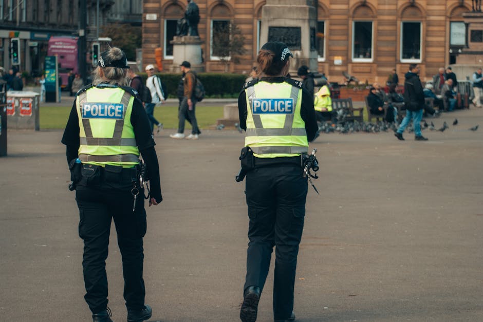 Two police officers in high-visibility vests patrol a busy urban park area, ensuring public safety.
